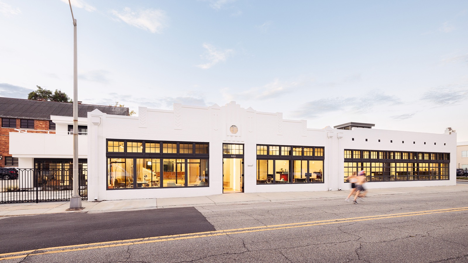 The Lewis Communications building in Mobile, Alabama at dusk — a white painted-brick storefront with black steel windows glowing warm, two runners passing on the sidewalk